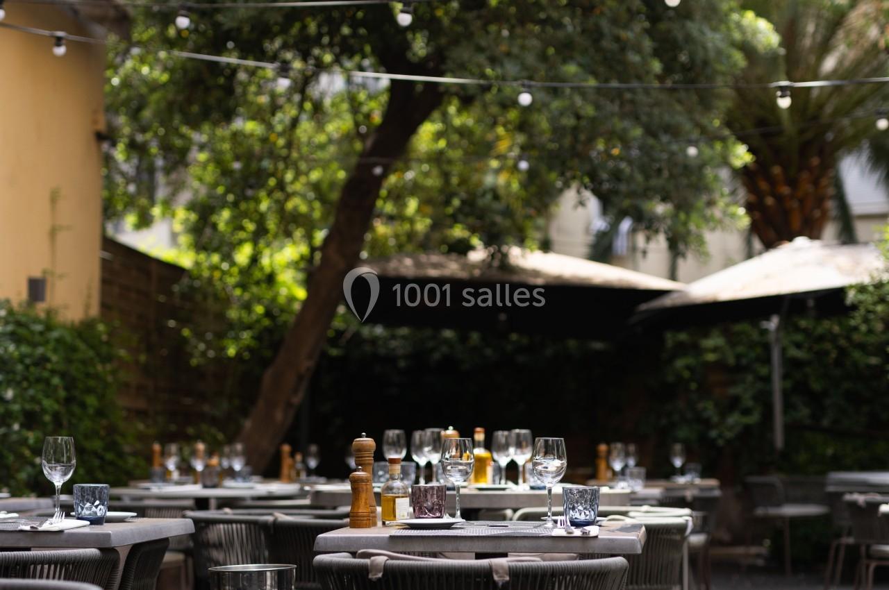 Terrasse extérieure avec tables dressées, chaises en rotin, guirlandes lumineuses et arbres en arrière-plan.