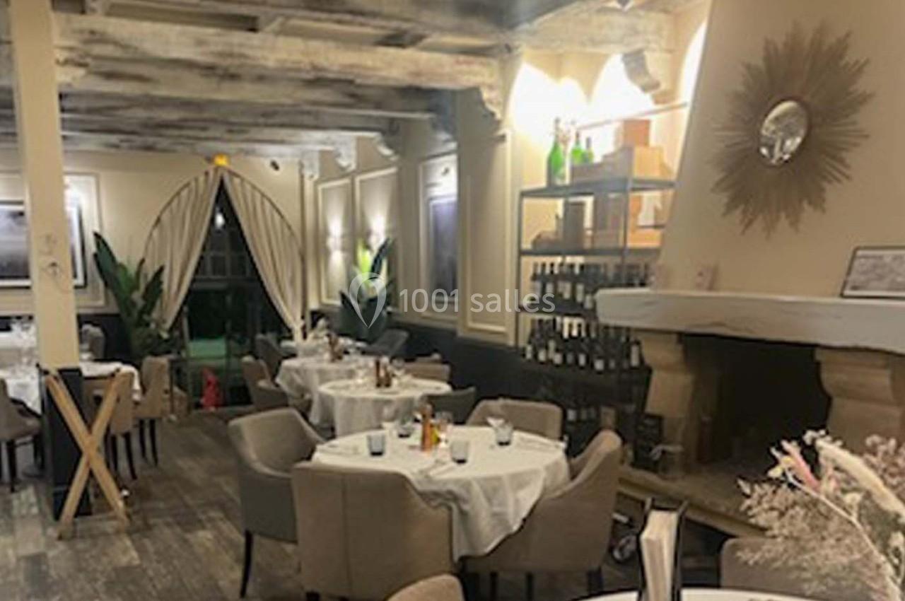 Salle de restaurant élégante avec tables dressées, cheminée, étagères à vin et poutres apparentes au plafond.