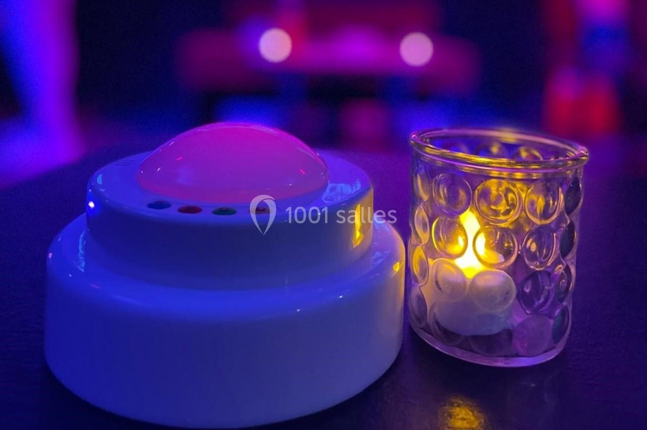 Une lumière LED blanche et une bougie dans un photophore en verre sur une table dans une ambiance tamisée.