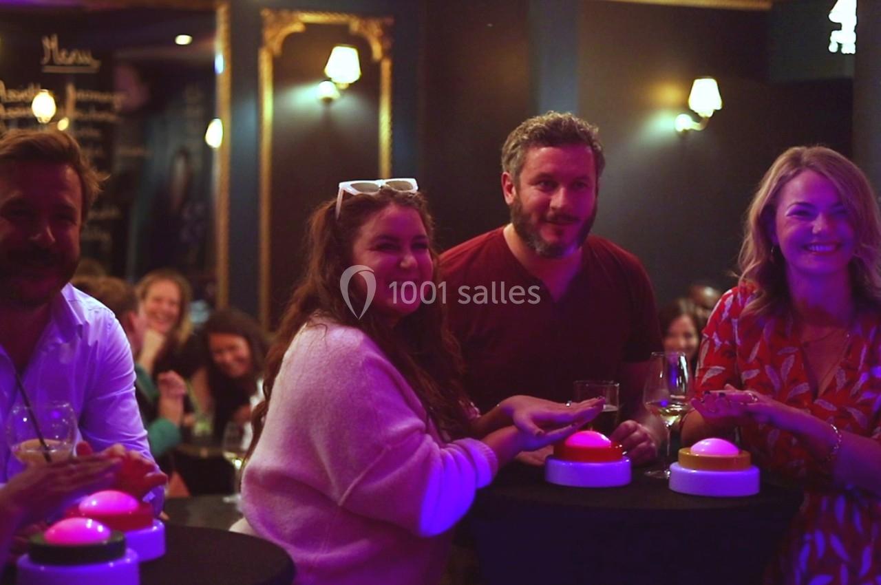 Des personnes souriantes autour de tables hautes avec des buzzers lumineux, dans une ambiance conviviale et tamisée.