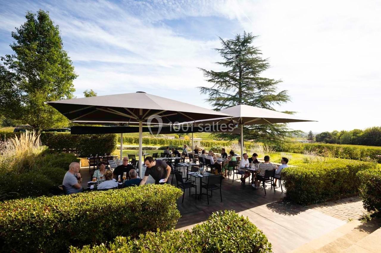 Terrasse extérieure d'un restaurant avec des tables occupées, entourée de verdure et protégée par de grands parasols.