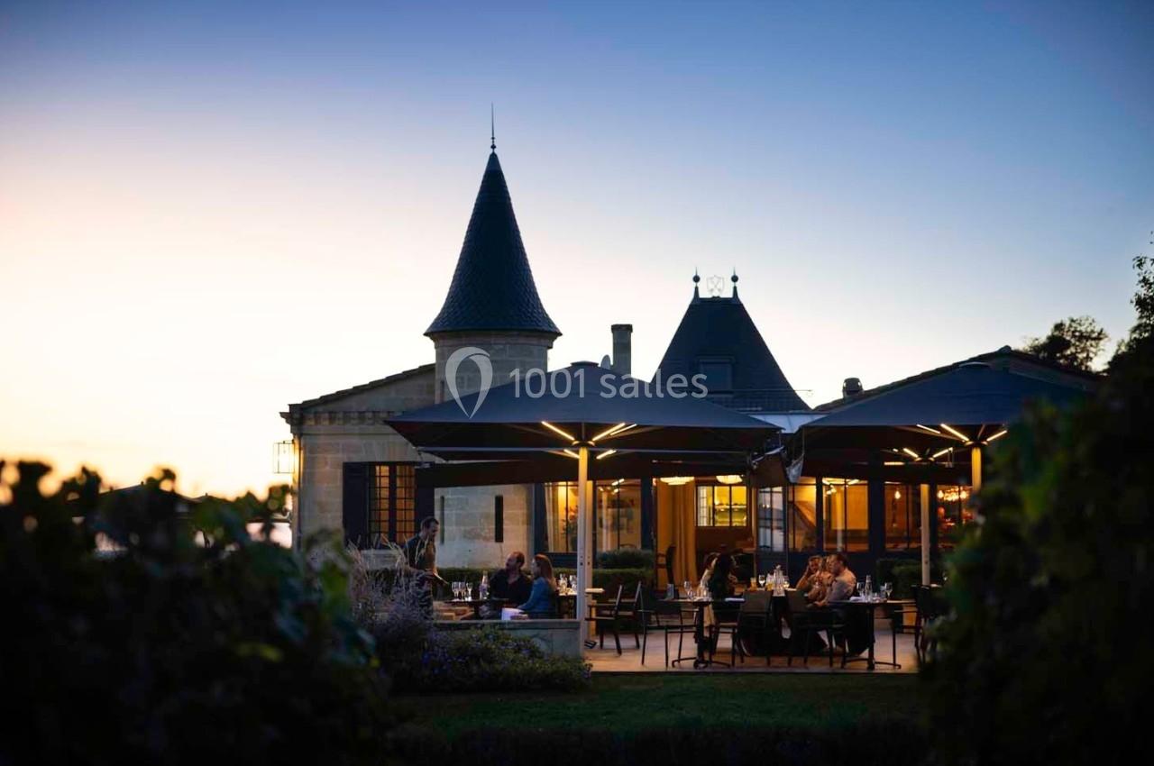Terrasse d'un restaurant éclairée au crépuscule, située devant un bâtiment de style château avec des tours.