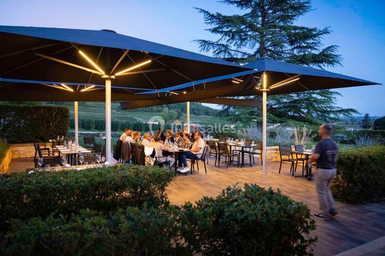 Terrasse d'un restaurant en soirée avec des convives attablés sous de grands parasols éclairés, entourée de verdure.