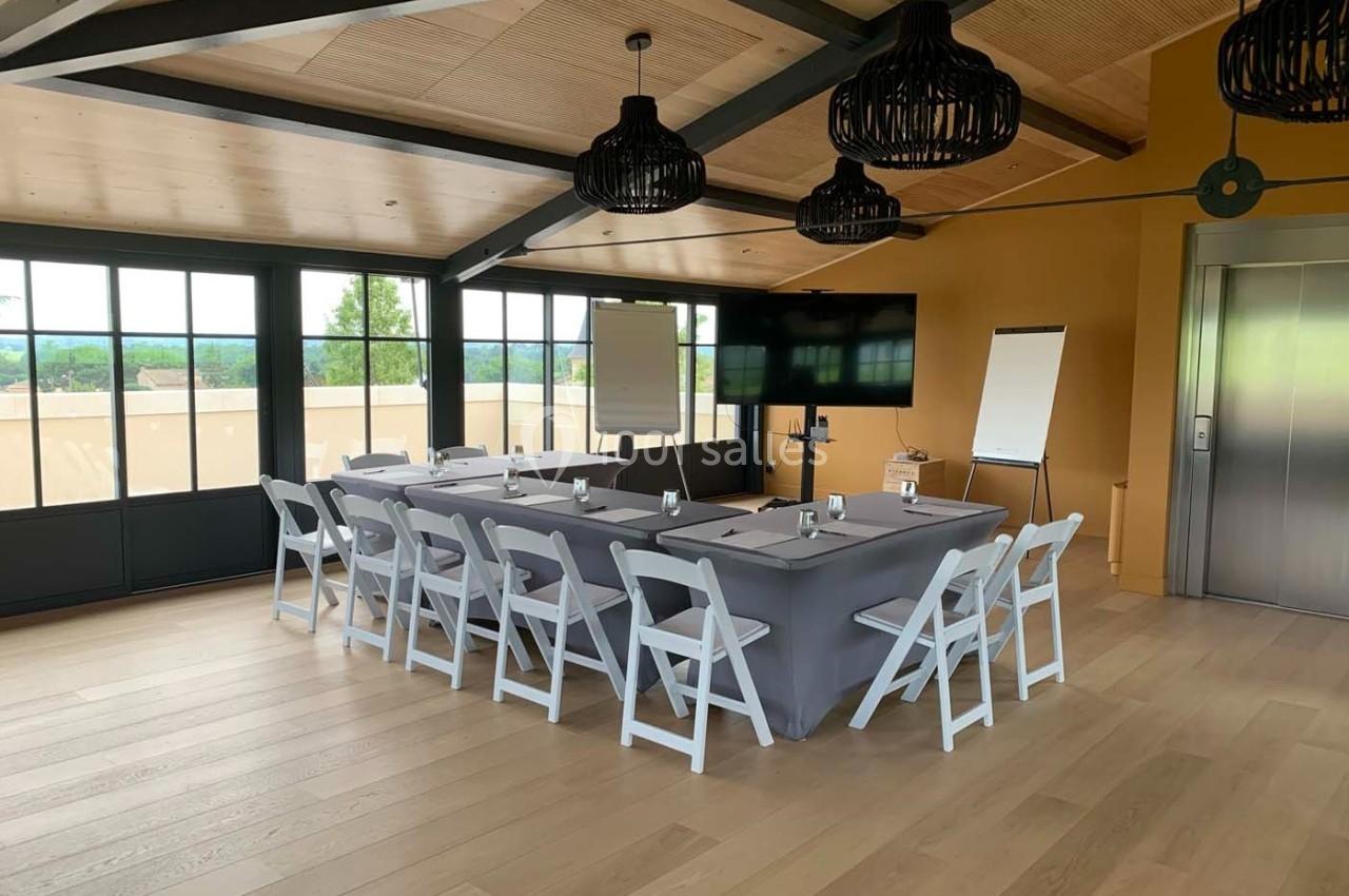 Salle de réunion lumineuse avec table rectangulaire, chaises blanches, tableau blanc et écran, vue sur l'extérieur.
