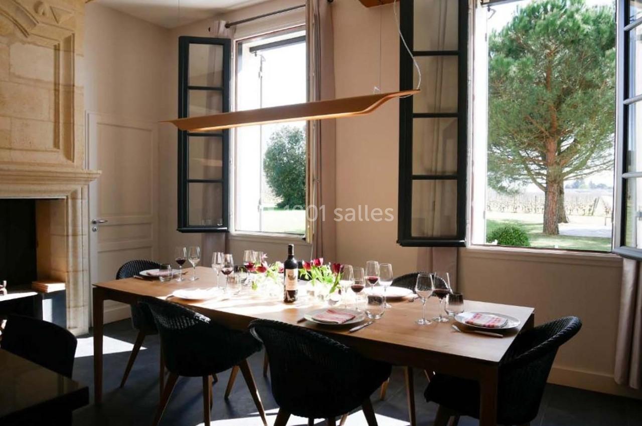 Salle à manger lumineuse avec une grande table en bois, des chaises noires, des fenêtres ouvertes et vue sur un arbre.