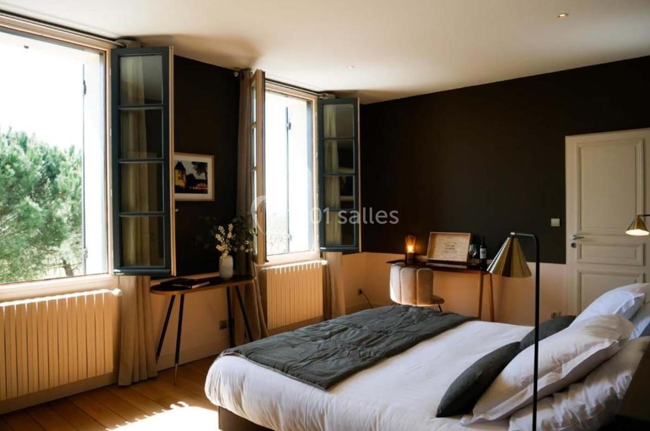 Chambre lumineuse avec lit double, bureau en bois, murs sombres et fenêtres ouvertes donnant sur des arbres.