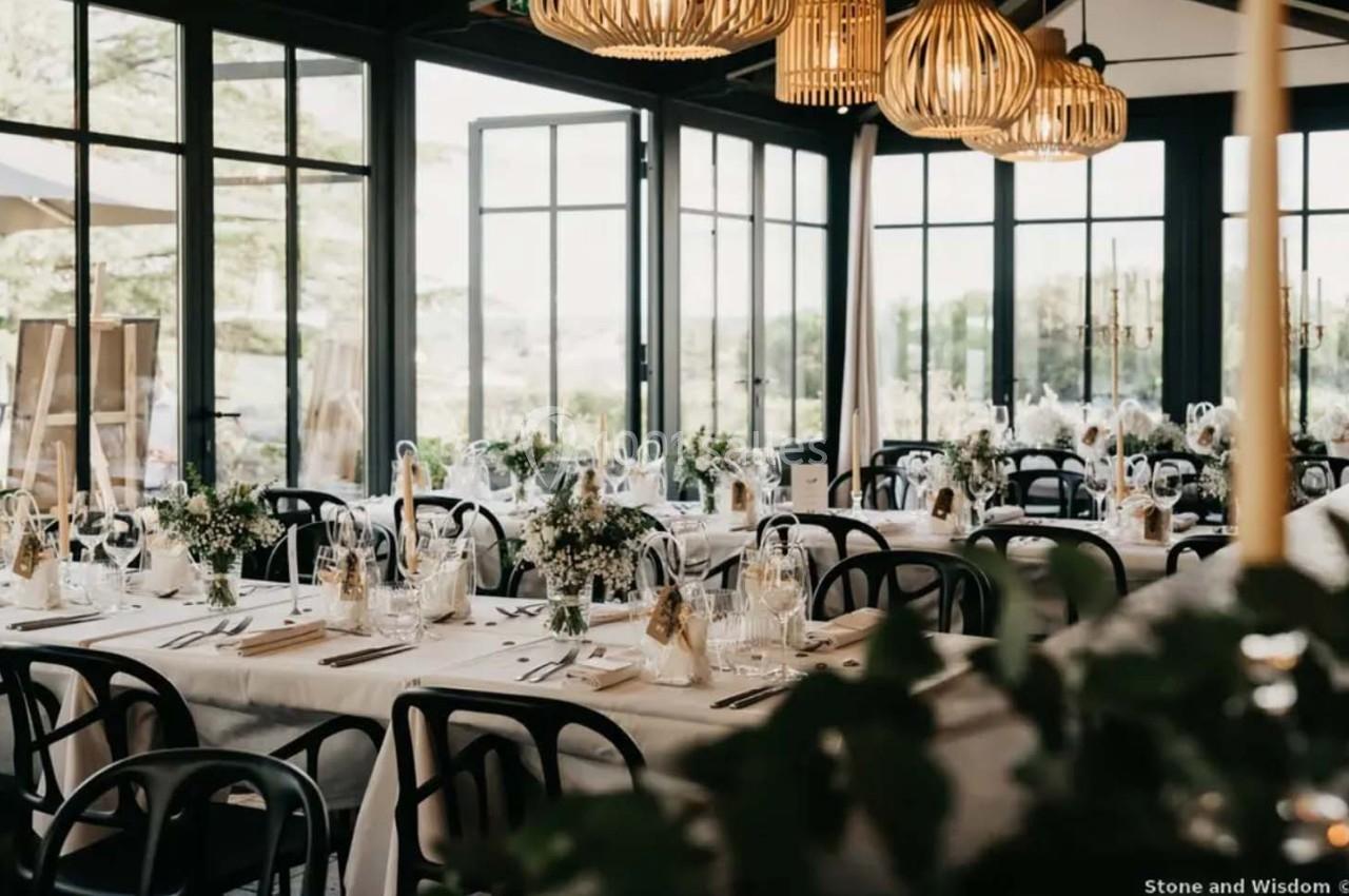 Salle de réception lumineuse avec tables décorées de fleurs et de bougies, entourée de grandes baies vitrées.
