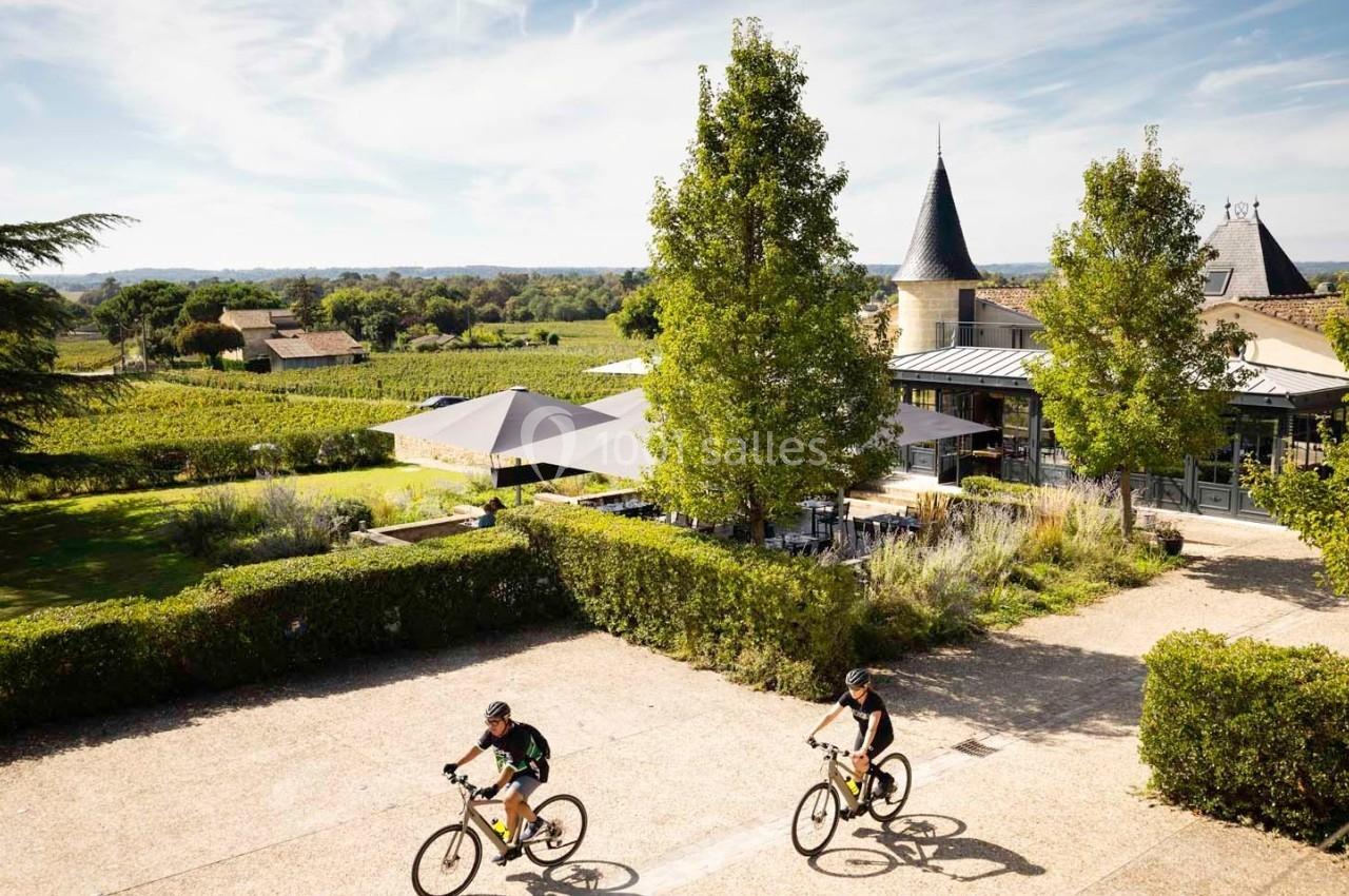 Deux cyclistes traversent une cour ensoleillée entourée de verdure, avec un château et des vignes en arrière-plan.