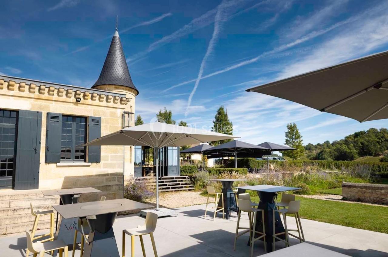 Terrasse extérieure avec tables, chaises et parasols, près d'un bâtiment en pierre avec une tour, sous un ciel dégagé.