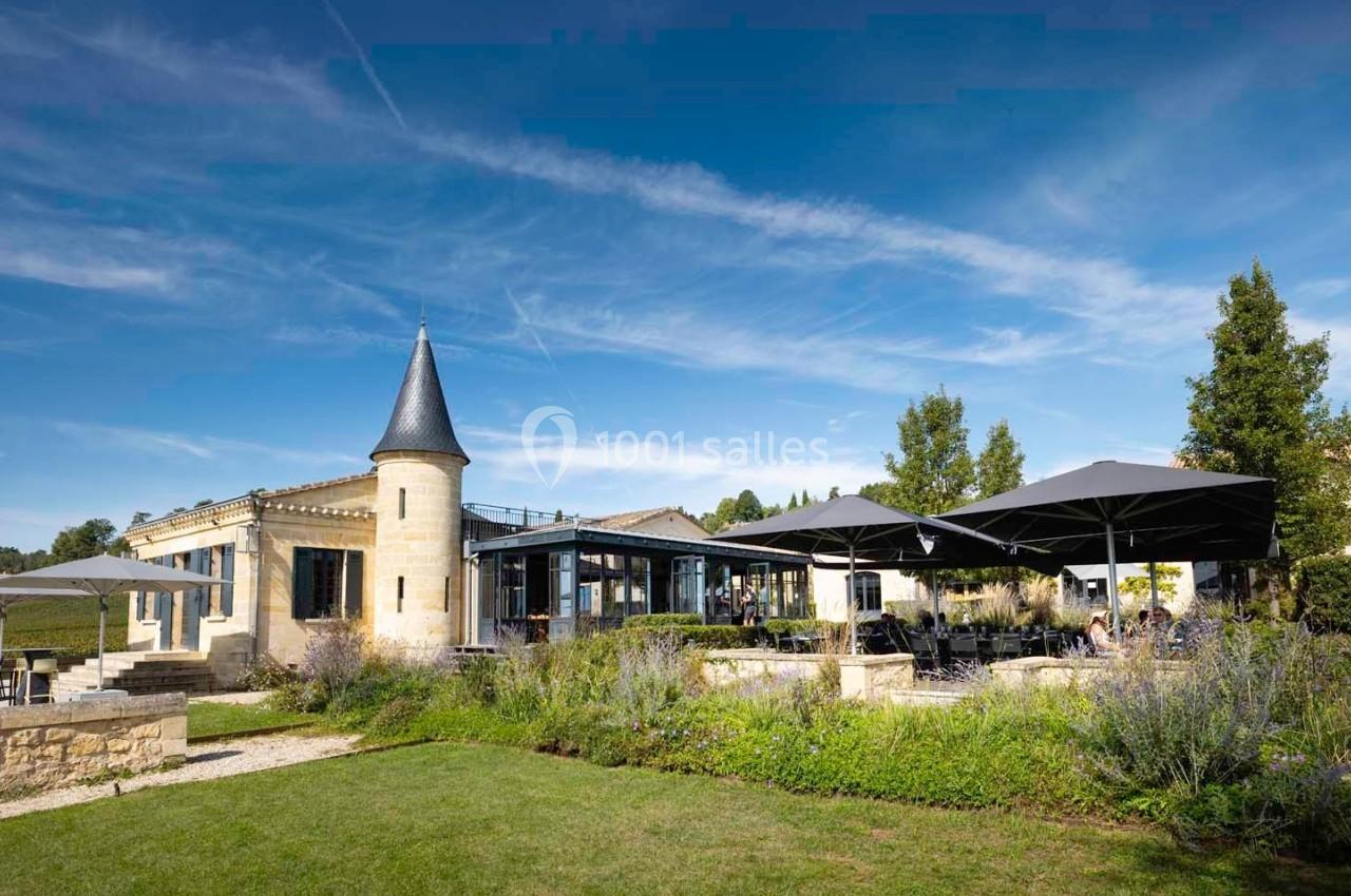 Bâtiment en pierre avec une tour ronde, terrasse ombragée par des parasols et jardin verdoyant sous un ciel bleu.