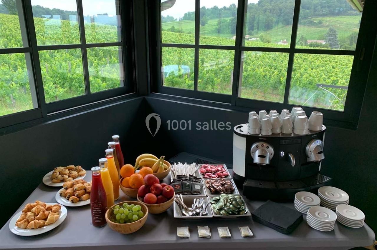 Table de petit-déjeuner avec viennoiseries, fruits, jus, café et vue sur un vignoble à travers de grandes fenêtres.
