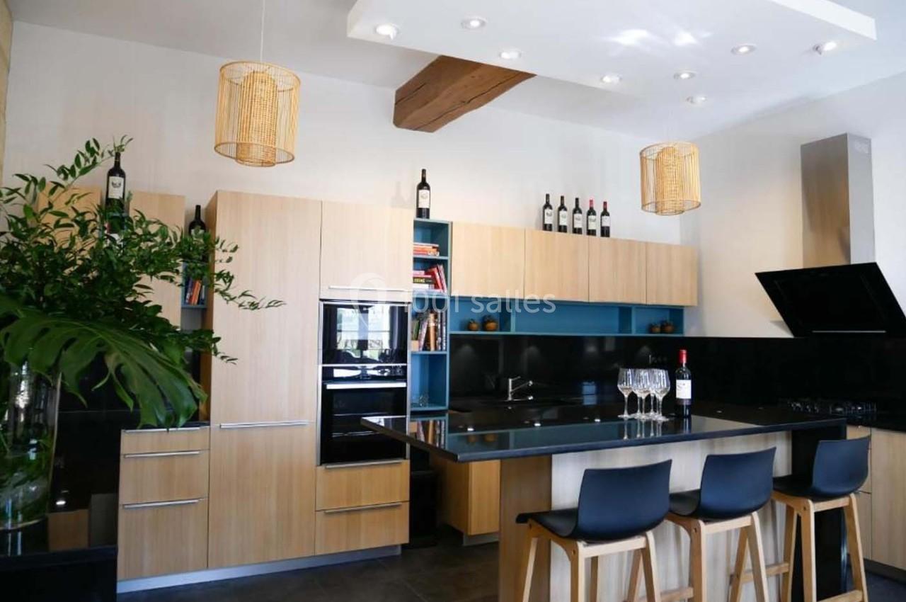 Cuisine moderne avec meubles en bois clair, îlot central, tabourets noirs et étagères décorées de bouteilles de vin.