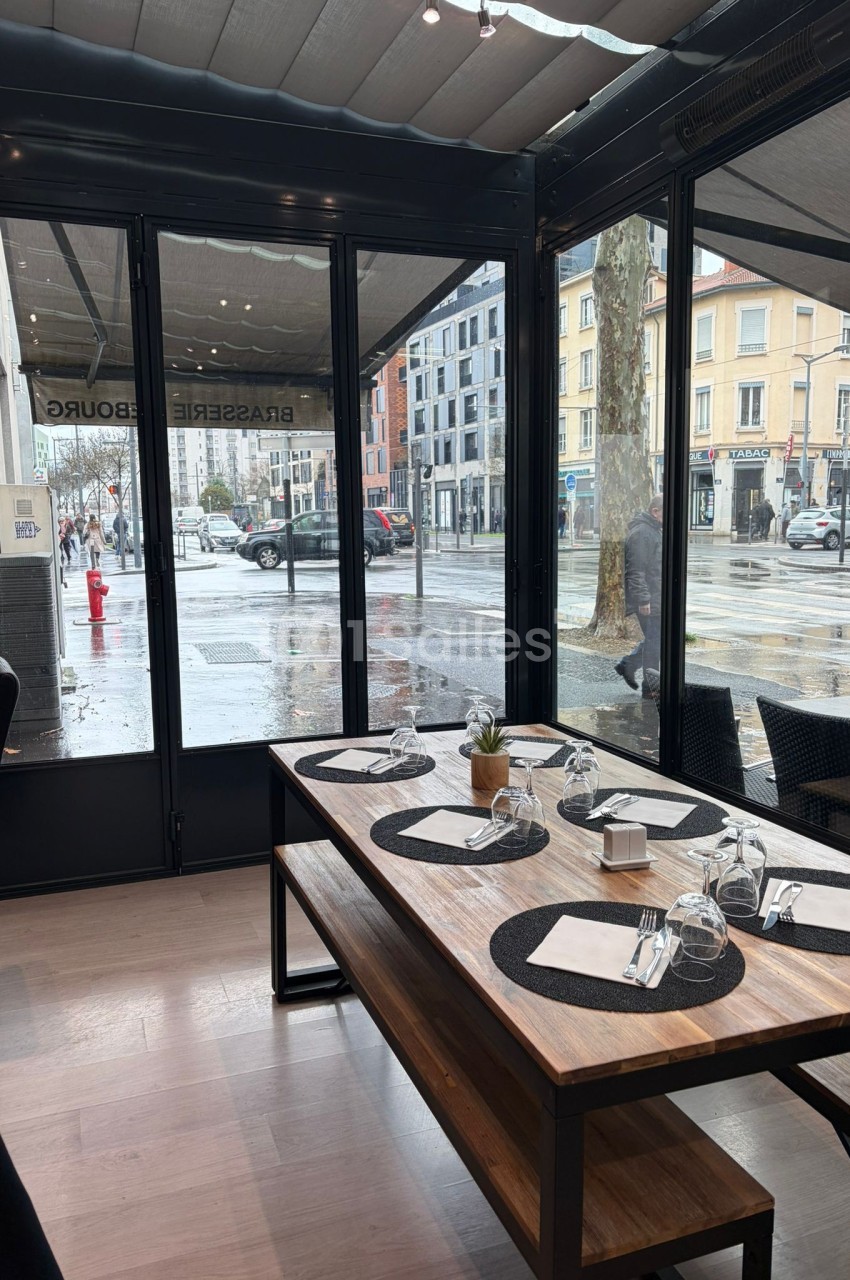 Table en bois dressée avec des sets noirs, des verres et des serviettes, située dans une véranda donnant sur une rue…