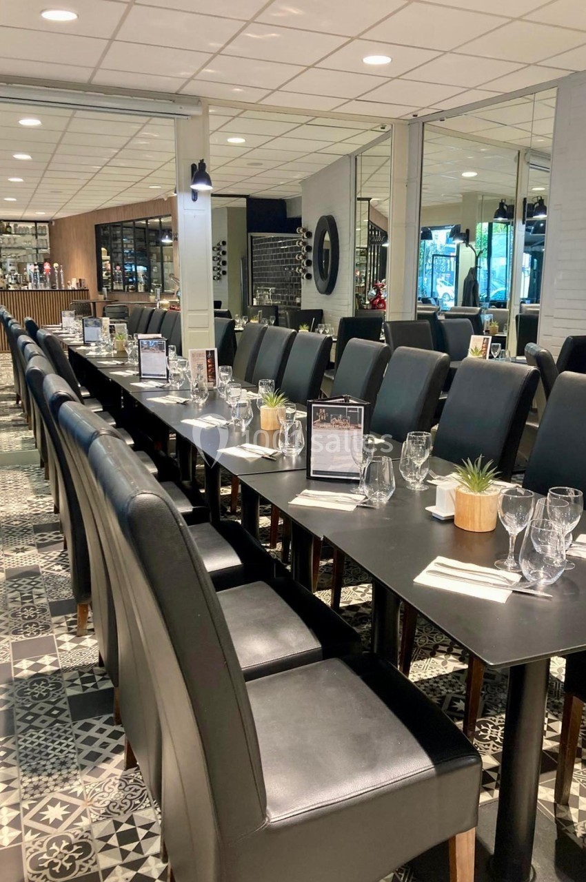 Salle de restaurant avec une longue table dressée, entourée de chaises noires, sur un sol à motifs géométriques.