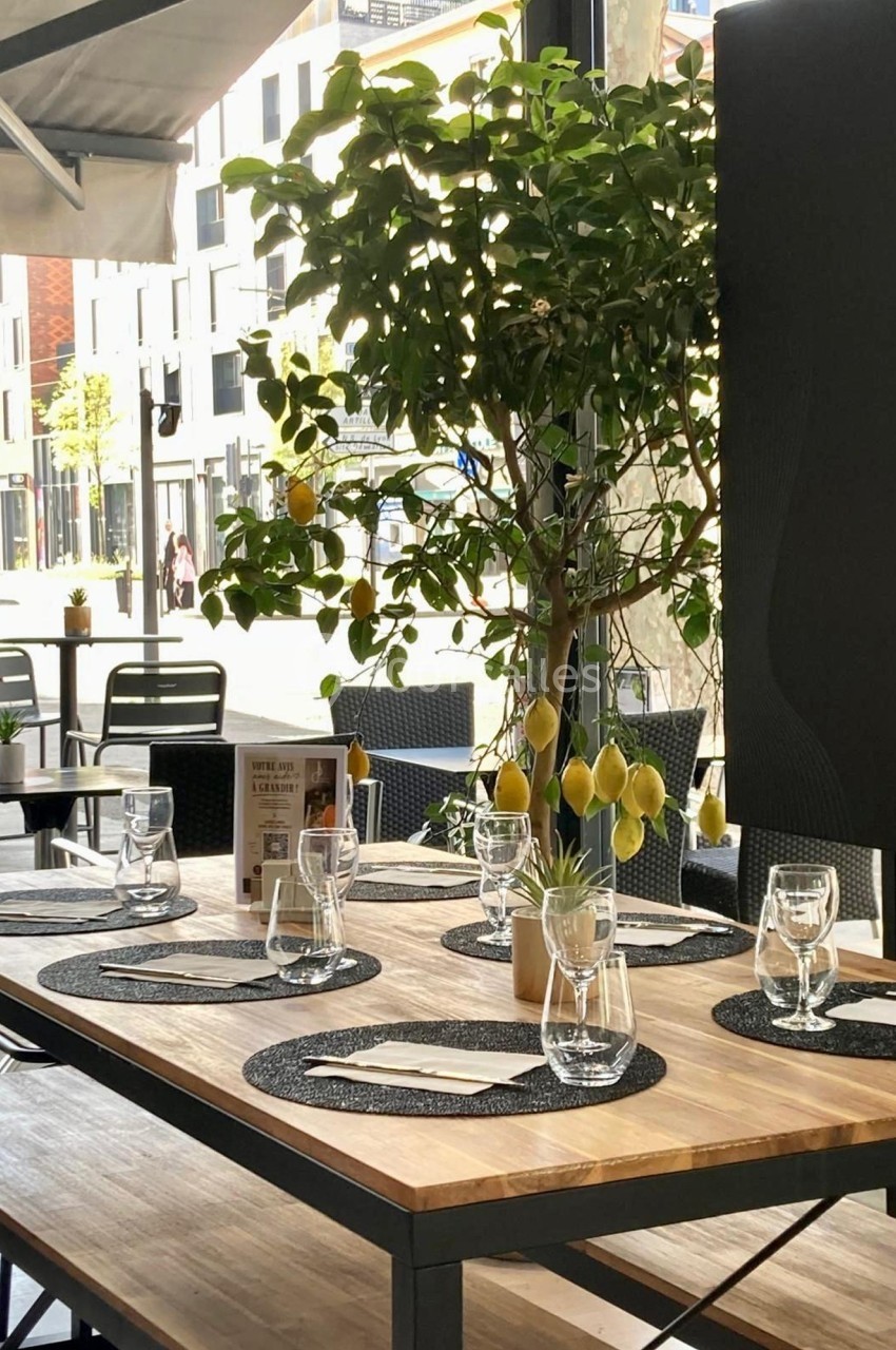 Table en bois dressée avec des sets noirs, verres et serviettes, décorée d'un citronnier en pot, en terrasse lumineuse.
