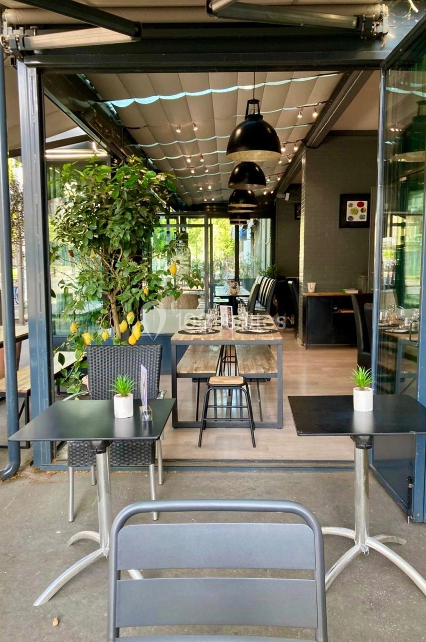 Salle de restaurant lumineuse avec tables dressées, plantes décoratives et grandes baies vitrées ouvertes.