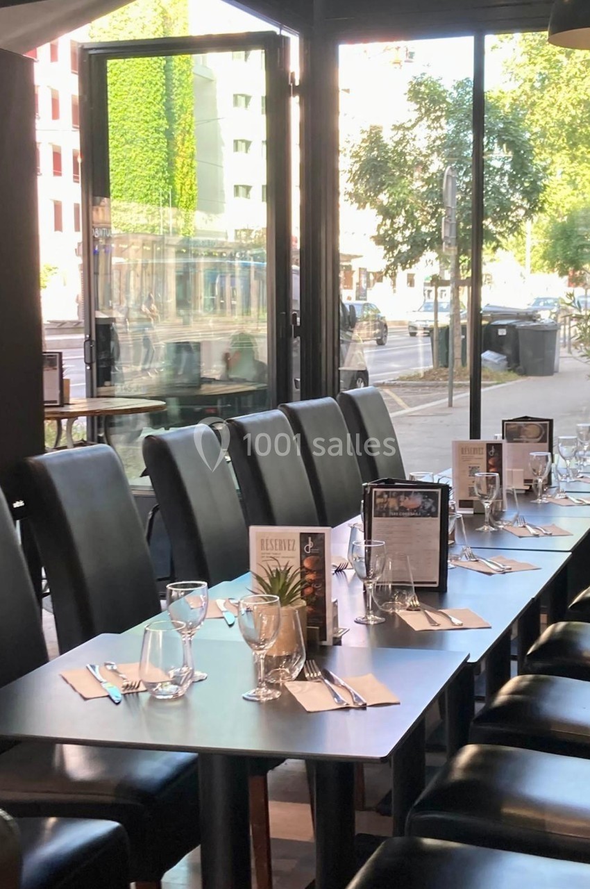 Salle de restaurant lumineuse avec tables dressées, chaises noires et vue sur une rue arborée à travers de grandes baies…