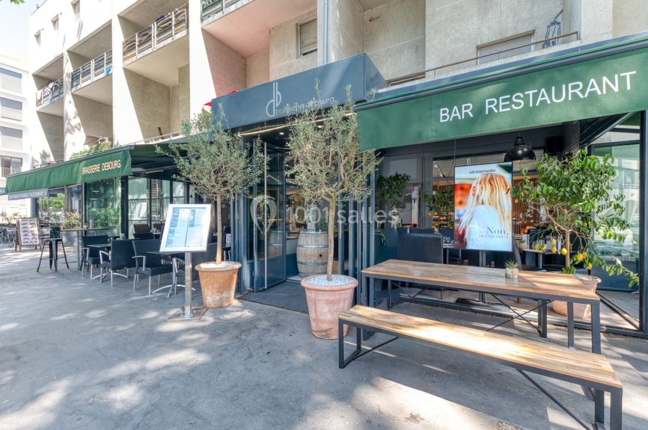 Façade d'un bar-restaurant avec terrasse, tables en bois, plantes en pot et enseignes vertes sous un auvent.