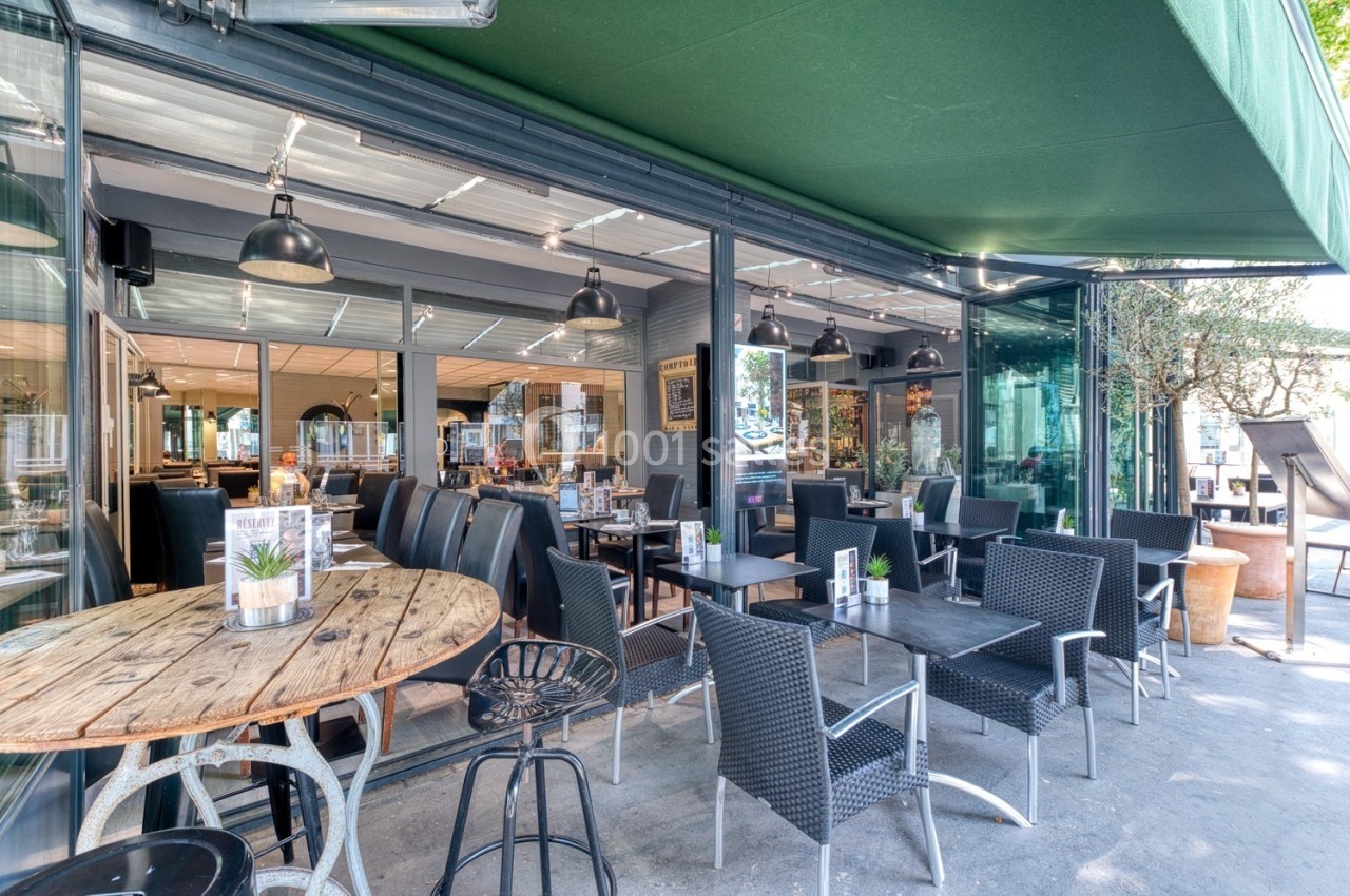 Terrasse d'un café-restaurant avec tables en bois et en osier sous un auvent vert, partiellement ouverte sur l'intérieur.