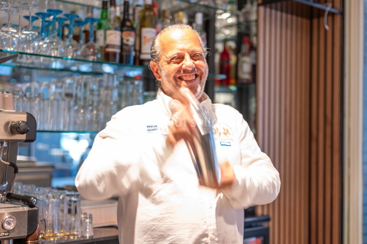 Un barman souriant secoue un shaker en métal devant un bar avec des étagères remplies de bouteilles et de verres.
