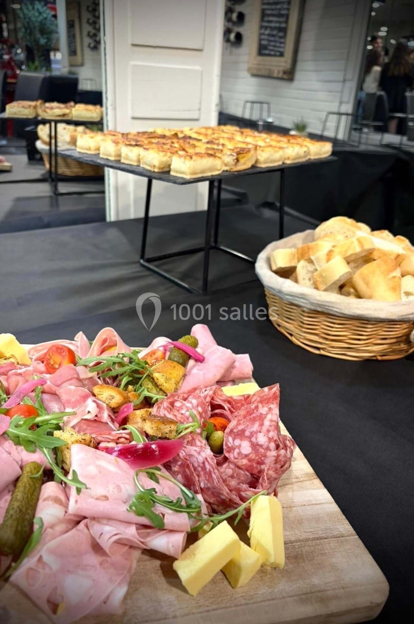 Plateau de charcuterie avec fromage et légumes en premier plan, paniers de pain et quiches en arrière-plan.