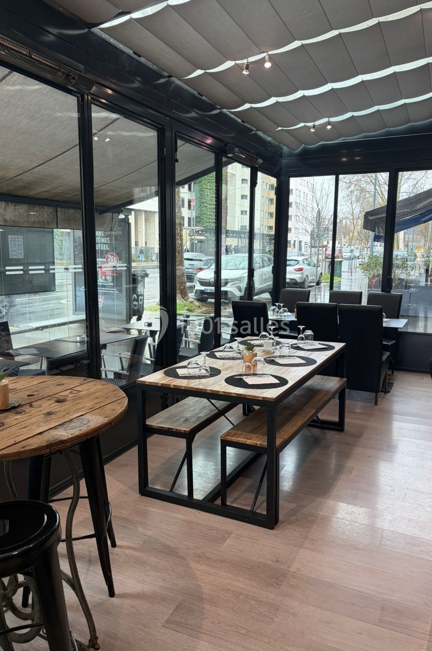 Salle de restaurant lumineuse avec grandes baies vitrées, tables en bois et chaises noires, vue sur l'extérieur urbain.