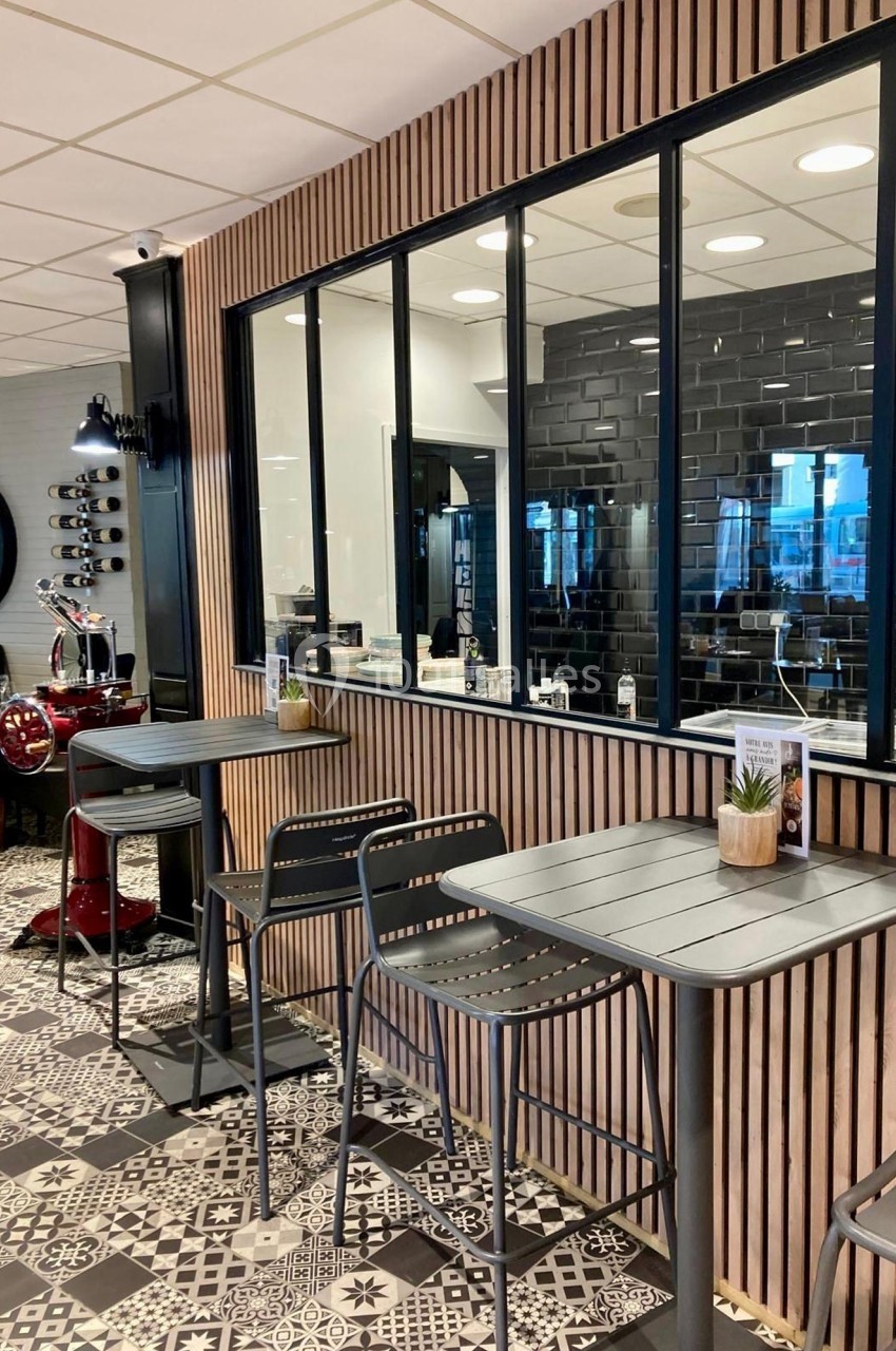 Espace intérieur avec tables hautes, chaises noires, cloison vitrée et carrelage à motifs en noir et blanc.