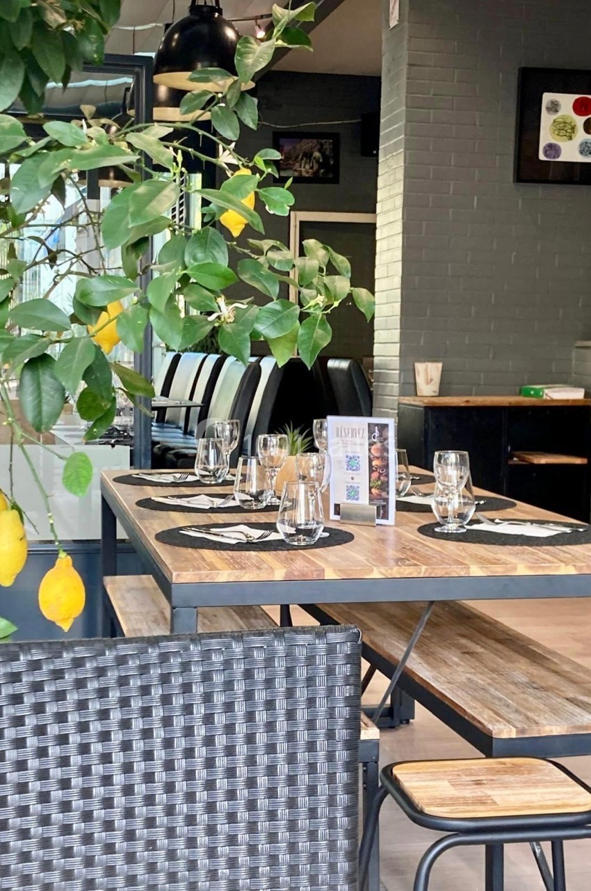 Salle de restaurant lumineuse avec tables en bois dressées, chaises noires et plante verte en premier plan.
