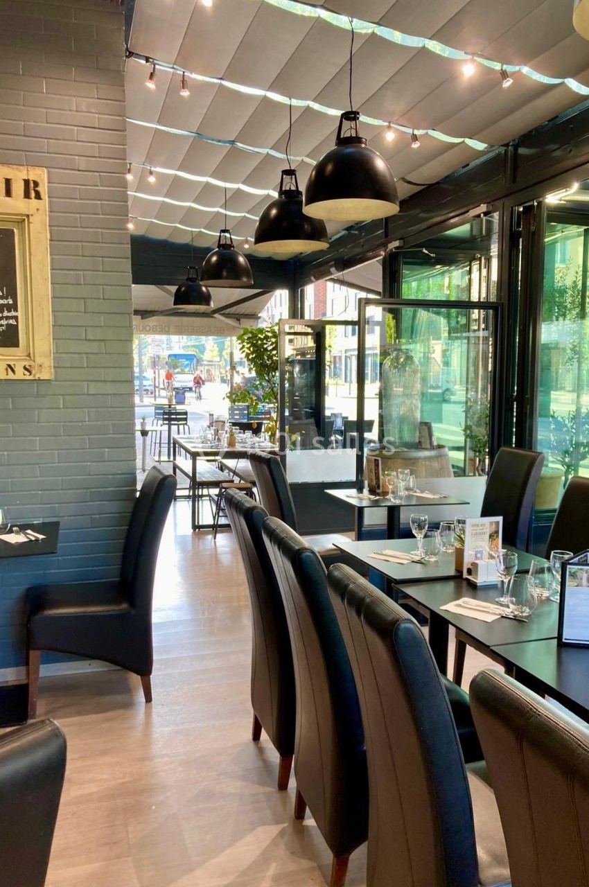 Salle de restaurant lumineuse avec tables dressées, chaises noires et grandes baies vitrées donnant sur l'extérieur.