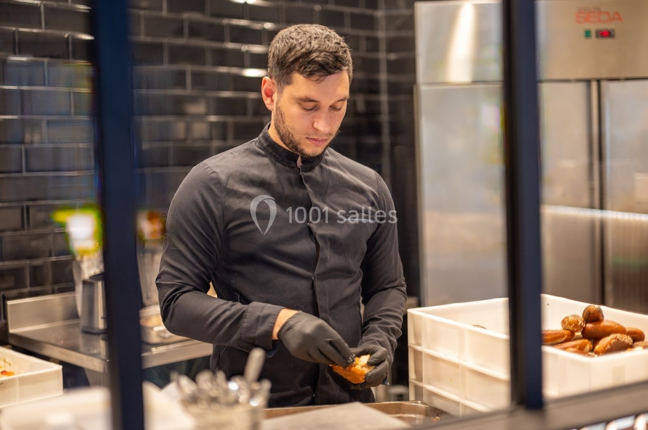 Un cuisinier en veste noire prépare des aliments dans une cuisine professionnelle avec des murs en carrelage noir.