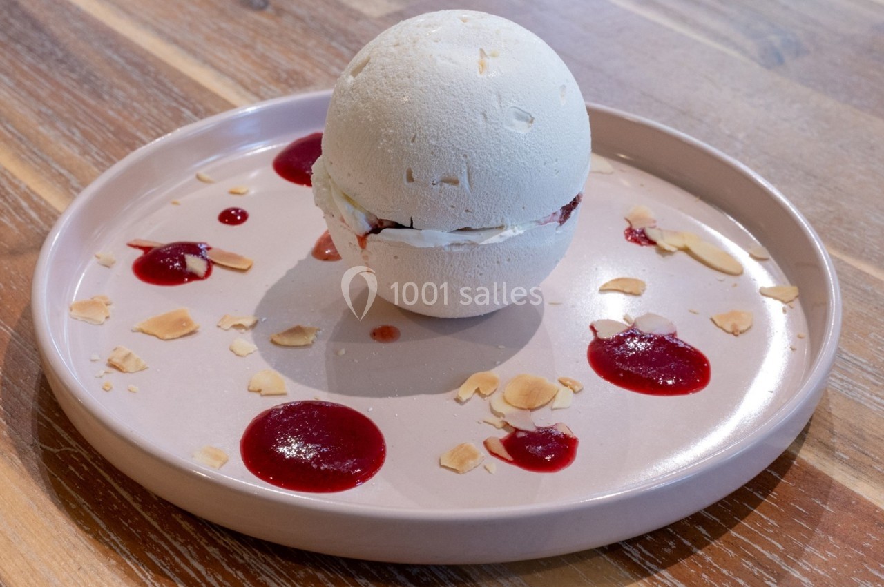 Dessert sphérique en meringue garni de crème et fruits rouges, entouré de coulis et éclats d'amandes sur une assiette.