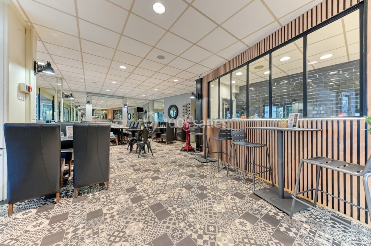 Salle de restaurant moderne avec mobilier noir, sol à motifs géométriques et grande baie vitrée donnant sur une cuisine.