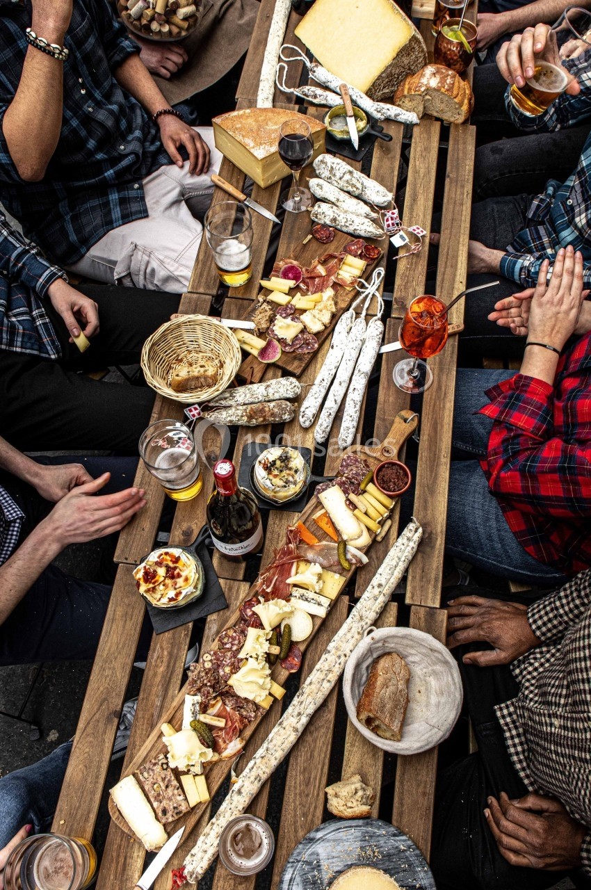 Table en bois garnie de charcuteries, fromages, pains et boissons, entourée de convives en chemises décontractées.