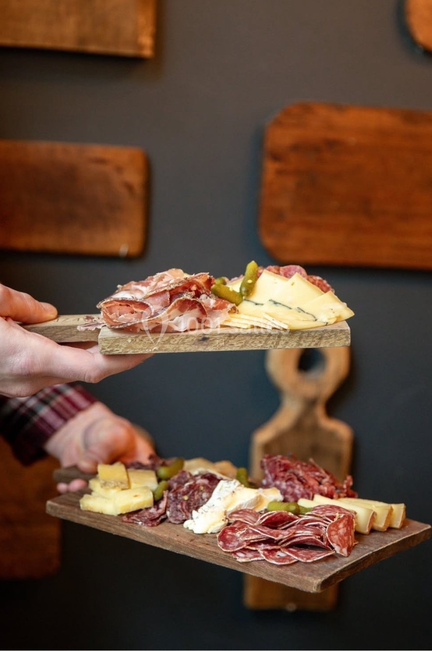 Deux planches en bois garnies de charcuterie, fromage et cornichons, tenues à la main devant un mur sombre.