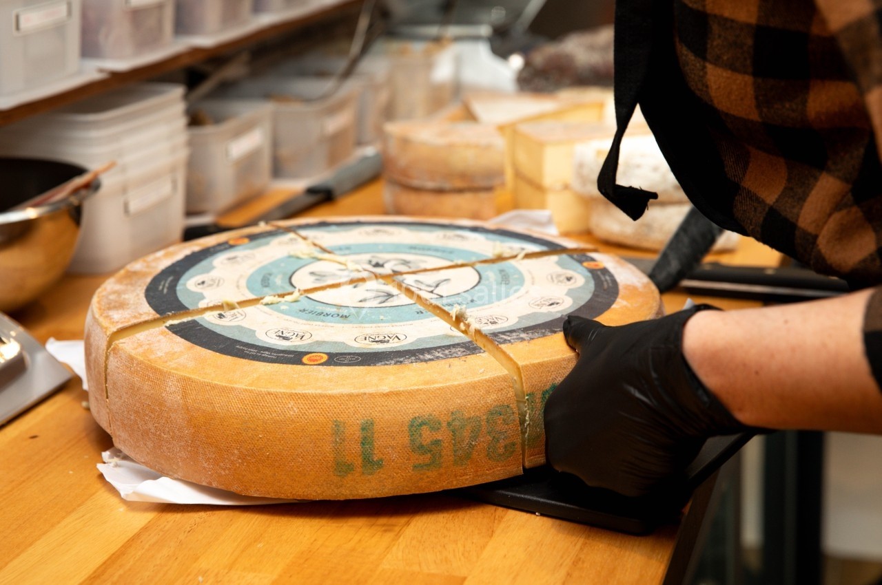 Une personne découpe une grande meule de fromage sur un plan de travail en bois, entourée d'ustensiles de cuisine.