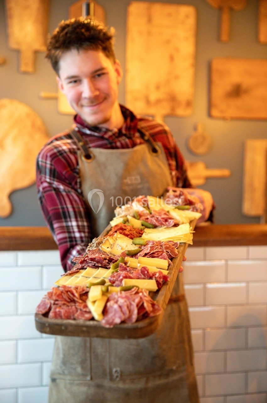 Un homme en tablier présente un grand plateau de charcuteries et fromages dans un décor rustique.