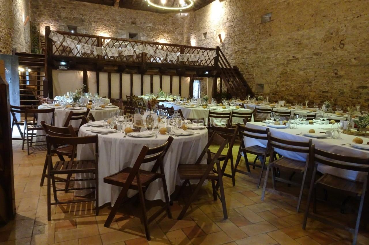 Salle en pierre avec des tables rondes dressées pour un repas, chaises en bois et mezzanine en bois visible.