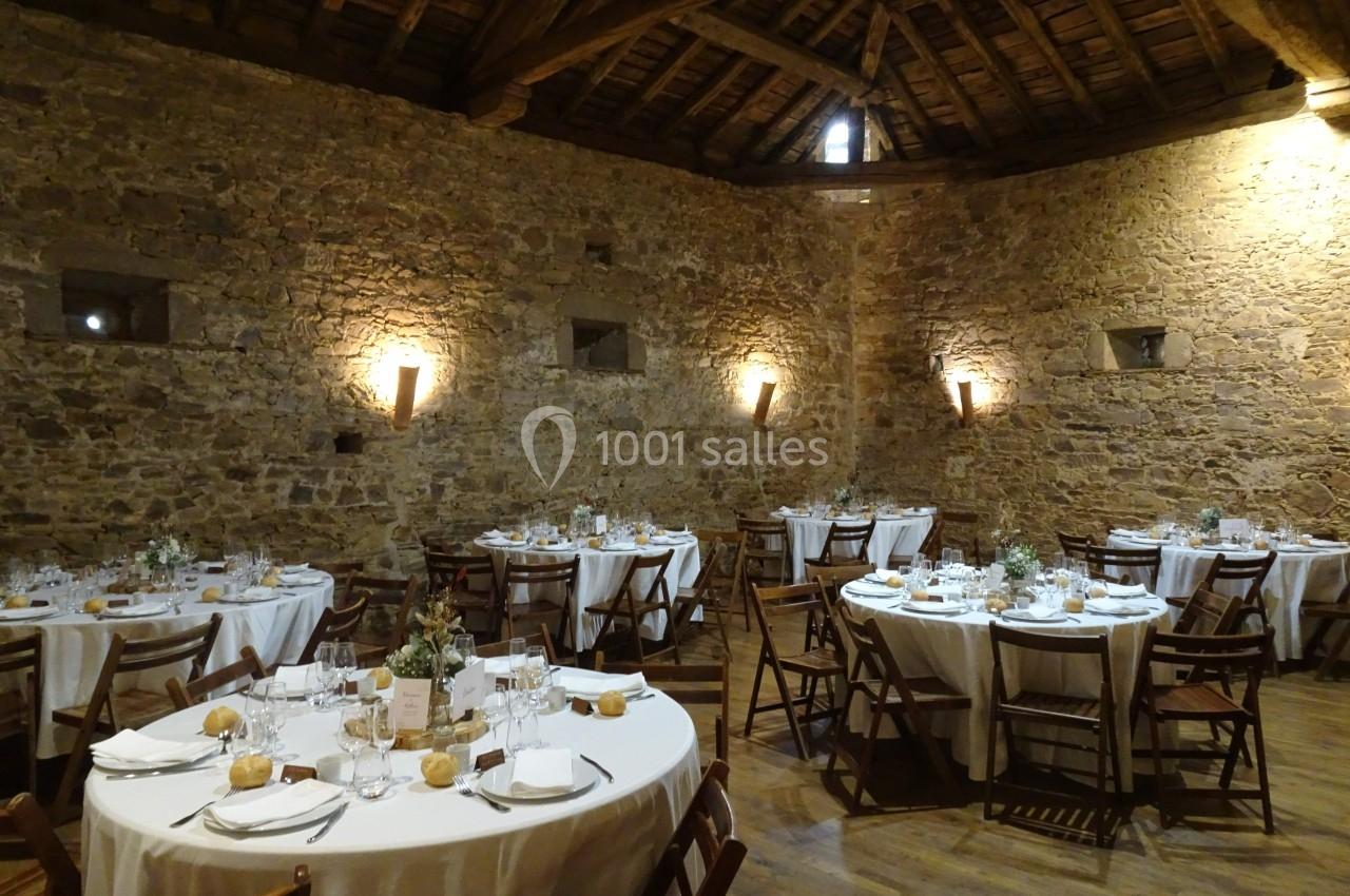 Salle en pierre avec tables rondes dressées pour un repas, éclairée par des appliques murales et une lumière naturelle.