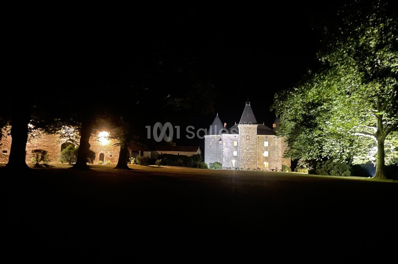 Château éclairé de nuit entouré d'arbres, avec une lumière illuminant une façade et une cour sombre au premier plan.