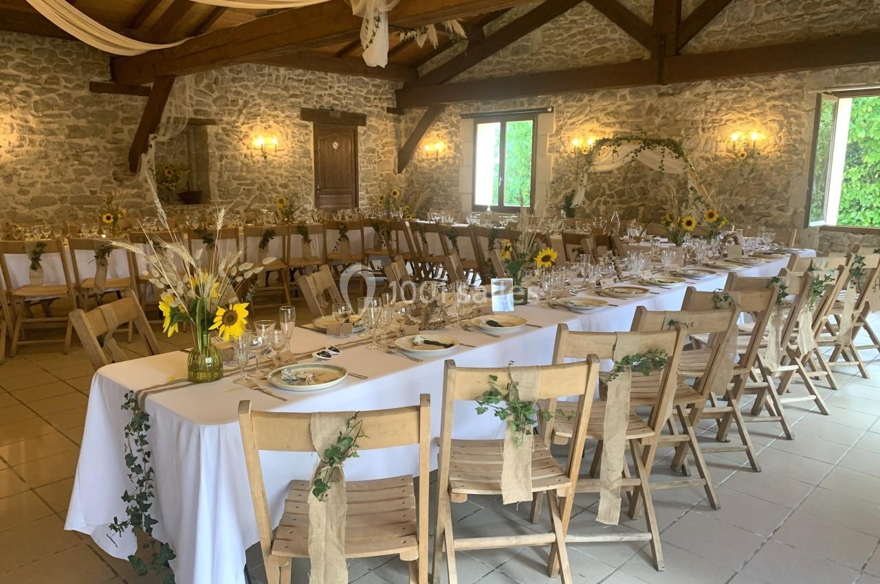 Salle de réception rustique décorée pour un événement, avec tables alignées, nappes blanches et ornements floraux.