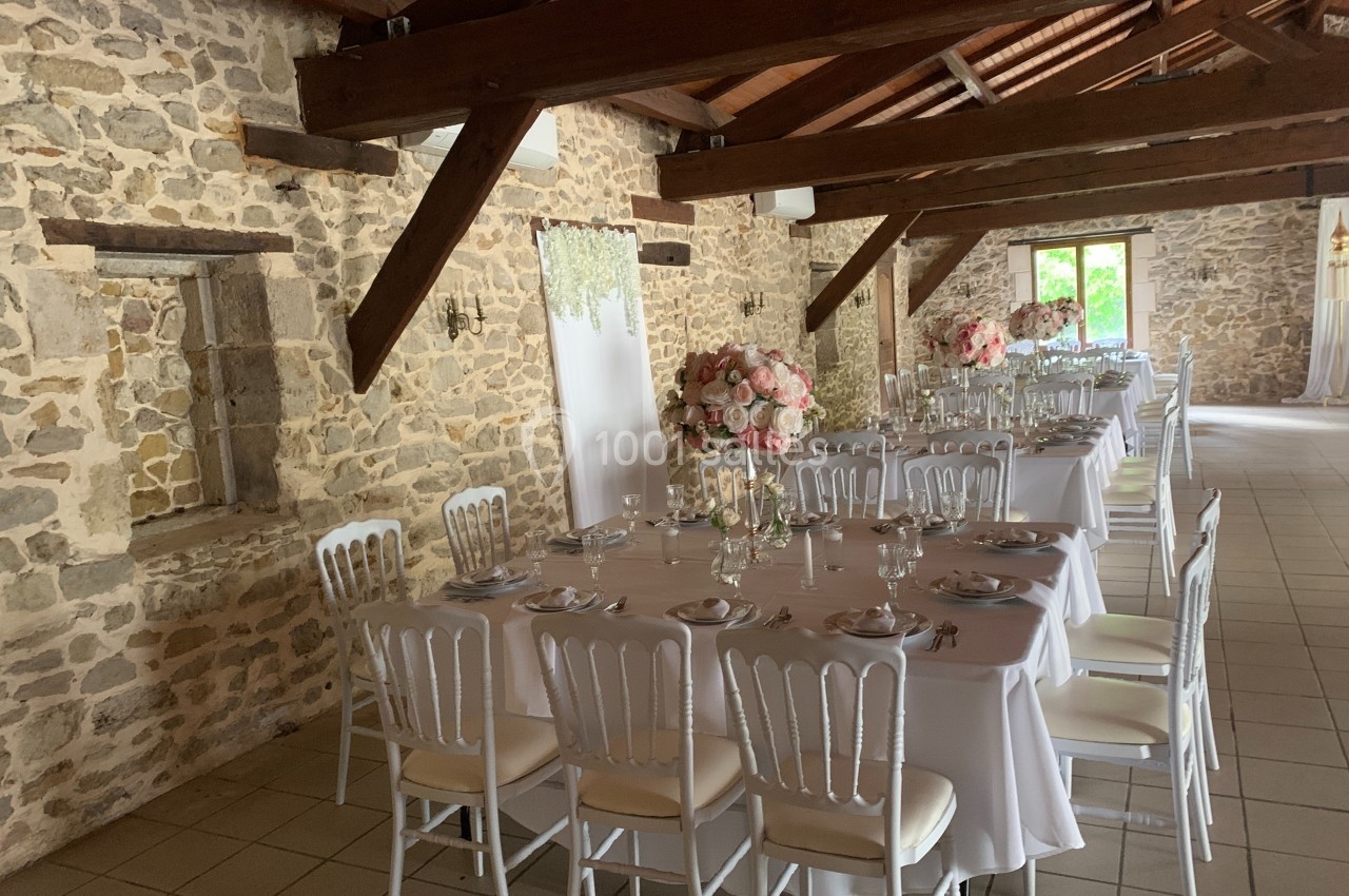 Salle en pierres apparentes avec tables dressées, nappes blanches, chaises élégantes et décorations florales roses.