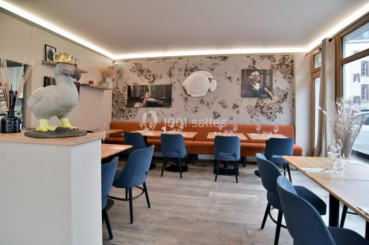 Salle de restaurant moderne avec tables en bois, chaises bleues, banquette orange et décoration murale artistique.