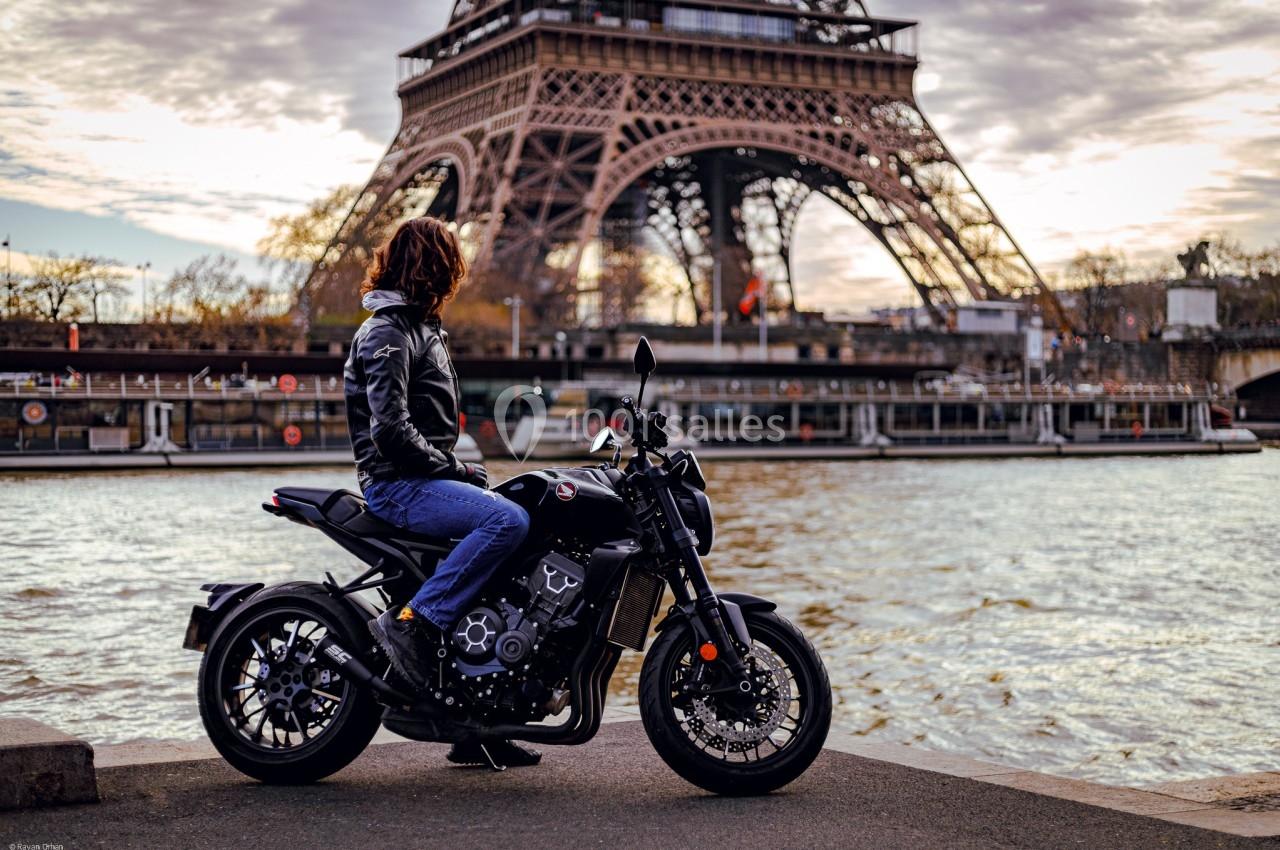 Une personne assise sur une moto noire près de la Seine, avec la tour Eiffel en arrière-plan par temps nuageux.