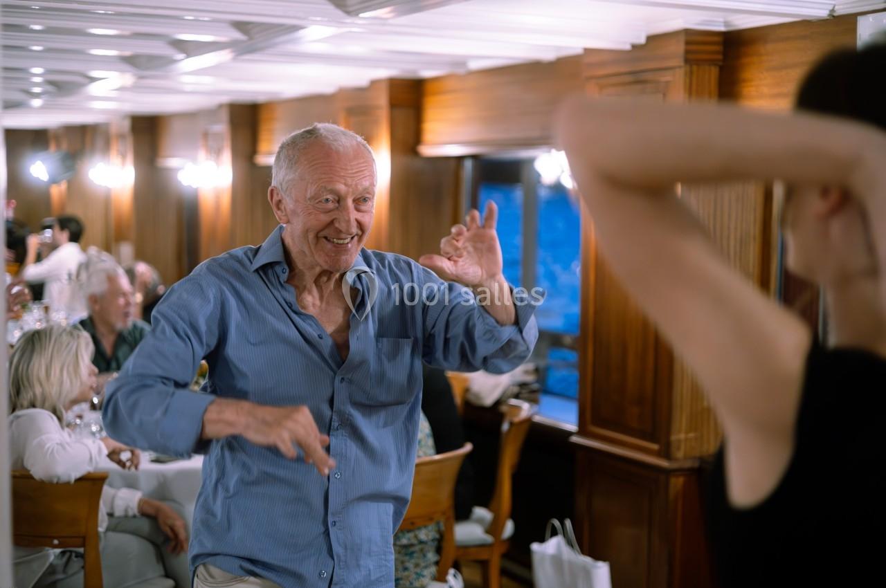 Un homme âgé danse joyeusement dans une salle en bois éclairée, entouré de personnes assises à des tables.