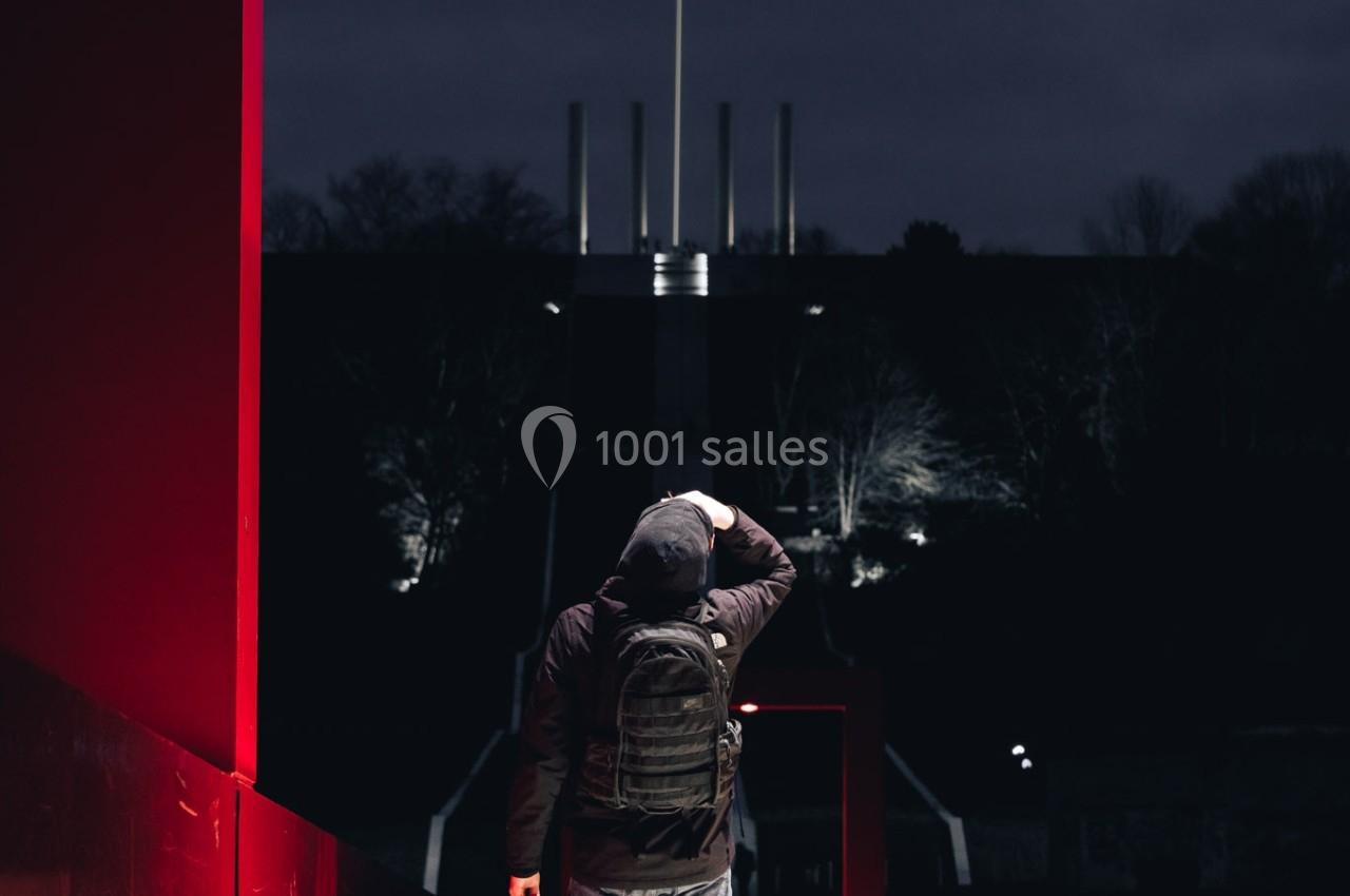 Un homme avec un sac à dos marche sur un pont éclairé par des lumières rouges, dans un environnement nocturne.