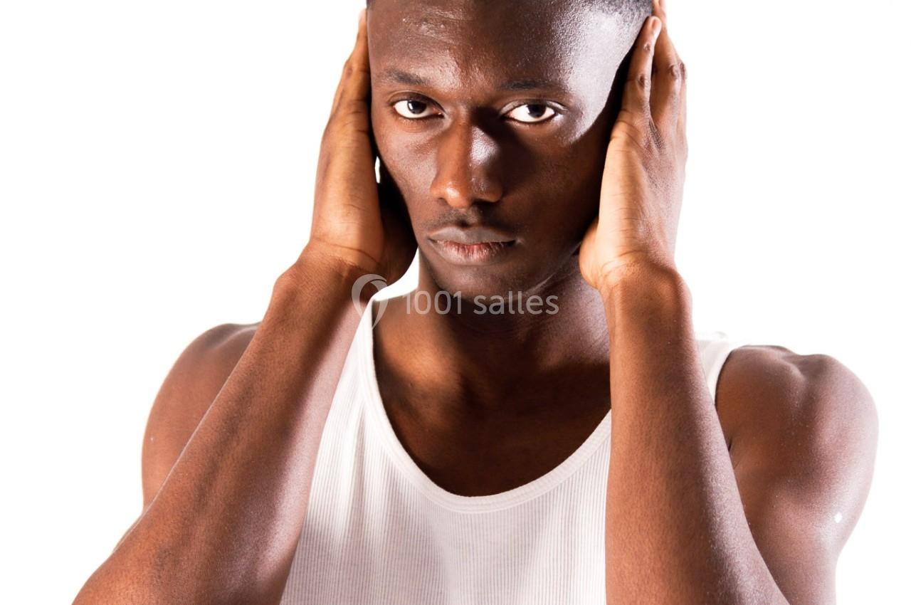 Un homme en débardeur blanc pose en tenant ses mains sur ses oreilles, sur un fond blanc.