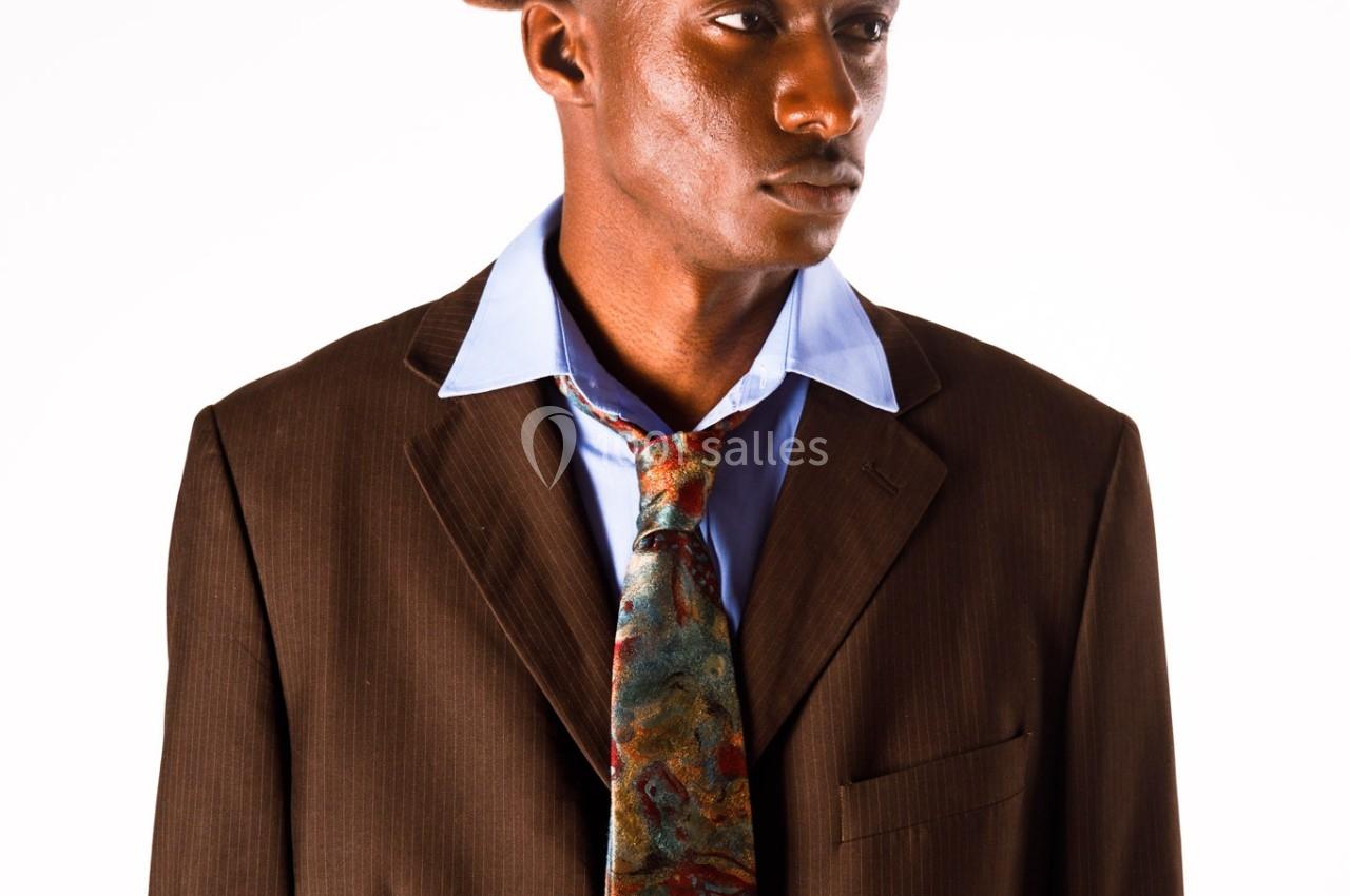 Homme portant un costume marron, une chemise bleue, une cravate colorée et un béret, sur fond blanc.