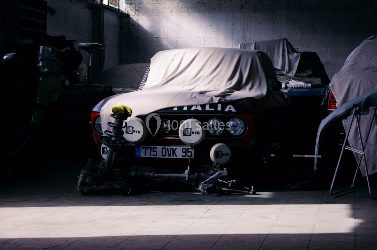 Voiture ancienne partiellement couverte dans un garage sombre, entourée de pièces détachées et d'autres véhicules.