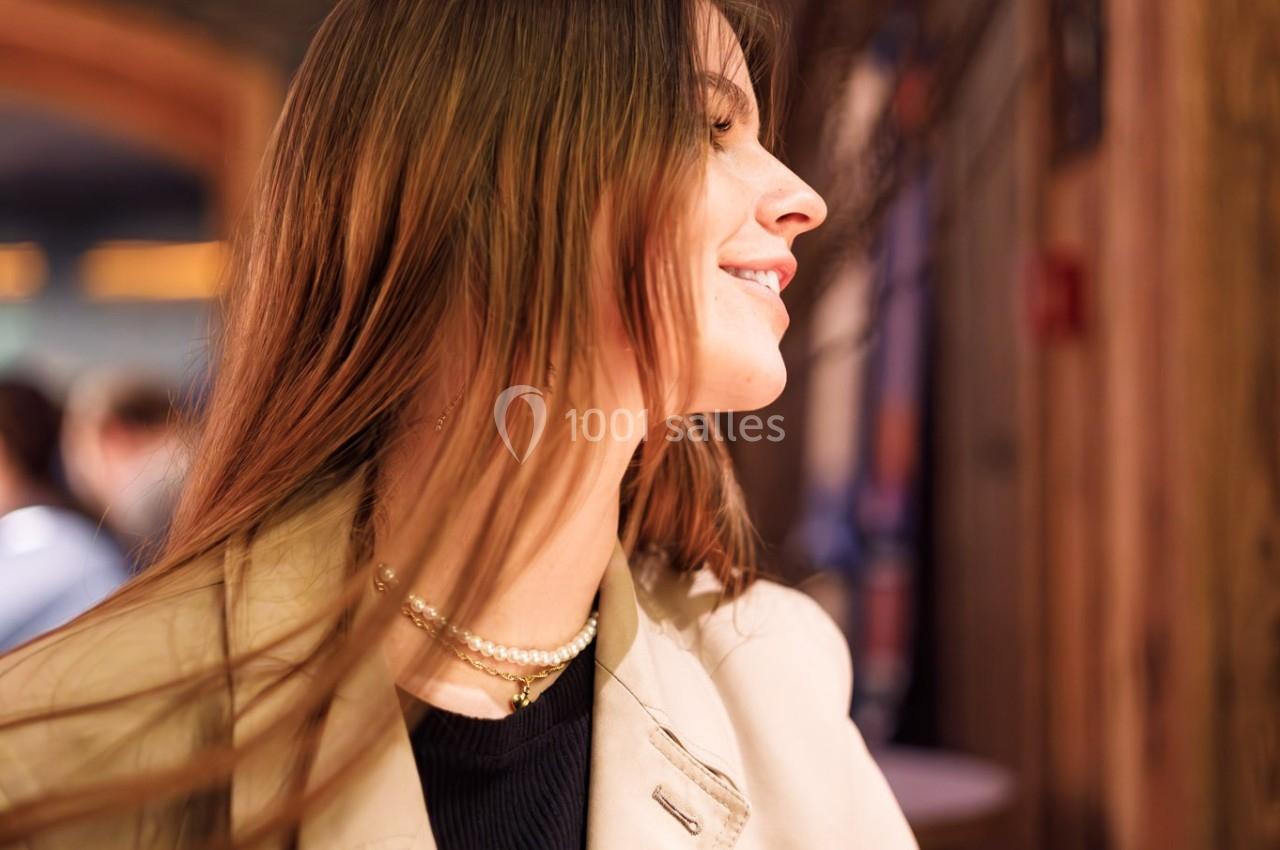Femme souriante portant un trench beige, assise dans un intérieur chaleureux avec des éléments en bois.