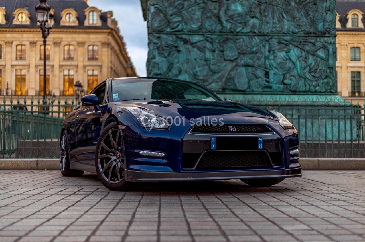 Voiture sportive bleue stationnée sur une place pavée devant un monument en bronze et des bâtiments historiques.